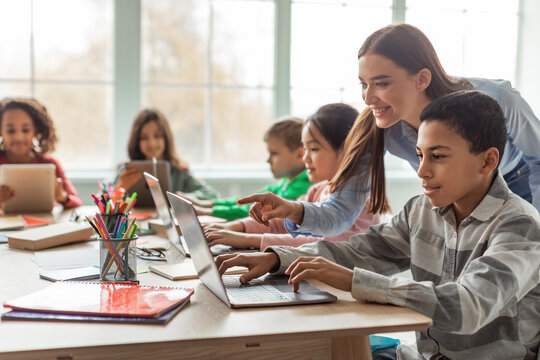 In a bright classroom, a teacher assists a group of diverse school children as they engage with laptops. The students, including an African American boy, explore online learning together. - Powered by Adobe