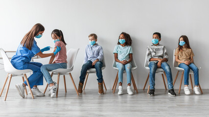 Nurse provides antiviral vaccine injections to a diverse group of children indoors. Kids are seated and waiting their turn in a clinic. The setting has a neutral gray wall backdrop.