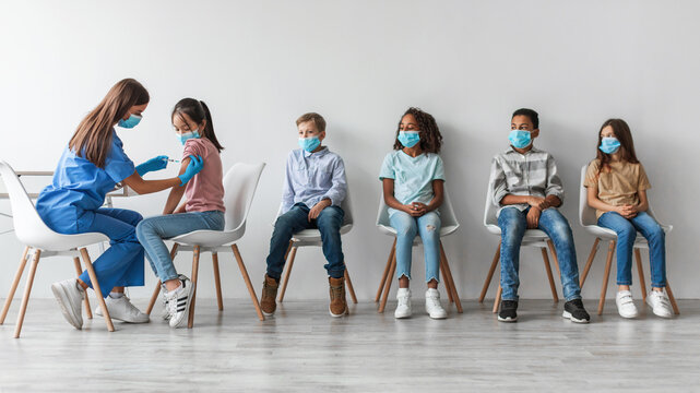 Nurse provides antiviral vaccine injections to a diverse group of children indoors. Kids are seated and waiting their turn in a clinic. The setting has a neutral gray wall backdrop.