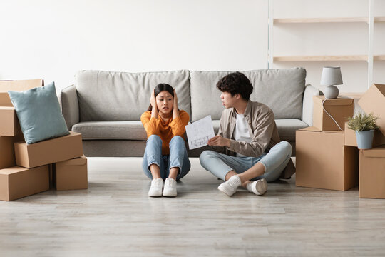 Stressed young Asian couple is having an argument about house rental or purchase documents in their new apartment. They sit among packed boxes on moving day.