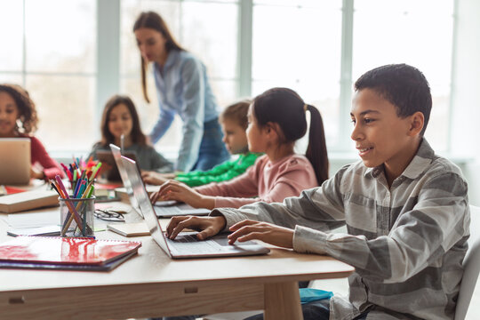 A cheerful Black schoolboy uses a laptop in a bright classroom. He is focused on online learning alongside diverse classmates, creating a collaborative and tech-friendly environment.