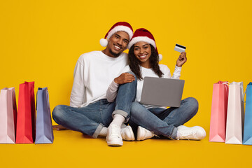 A happy couple sits on the floor, wearing Santa hats, smiling as they shop online. They have a laptop and a credit card ready, surrounded by vibrant shopping bags in various colors.
