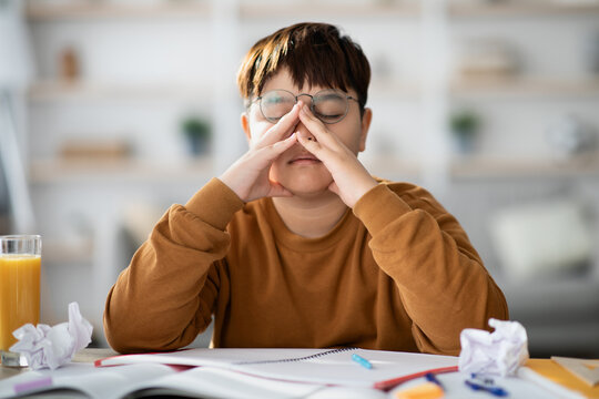 Overweight Japanese teenager is sitting at his desk doing a school project. He rubs his eyes in fatigue while surrounded by books and crumpled papers, seeking creative solutions.