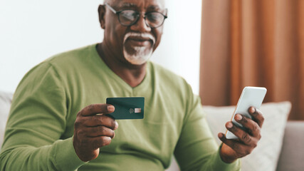 A senior man is seated on a couch in a comfortable living room, holding a smartphone and a credit card. He appears focused, engaging in a financial activity, reflecting modern technology.