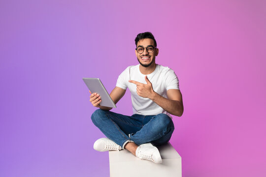 A handsome millennial Arab man sits cross-legged, checking a new app on his digital tablet. The scene is set against a neon light background, perfect for showcasing technology and creativity.