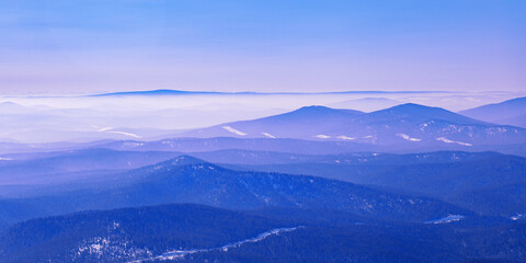 Panoramic view of mountain ridges covered morning mist and blue pink pastel colored sky banner. Distant peaks fade smoothly in horizon, creating depth effect under clear winter sky. Scenic landscape.