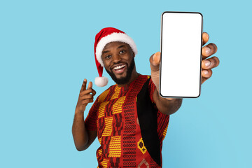 Holiday Offer. Excited Black Man In Traditional Costume And Santa Hat Pointing At Blank Smartphone In Hand, Cheerful African American Guy Demonstrating Mobile Phone With White Screen, Mockup