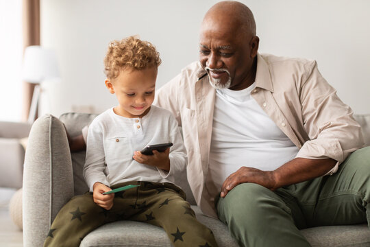 Black grandfather spends quality time with his little grandson as they explore a mobile game on a smartphone, enjoying each other's company in a comfortable living room.