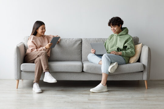 A young Asian couple sits comfortably on a couch in their living room, using a laptop and a tablet. They are engaged in online communication and enjoying their gadgets together.