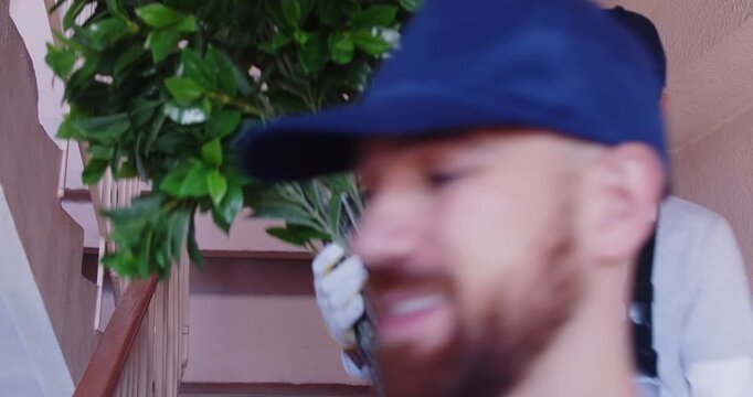 Two happy male home removals and delivery company workers in uniforms walking downstairs in hall carrying cardboard box and house plant. Moving service concept