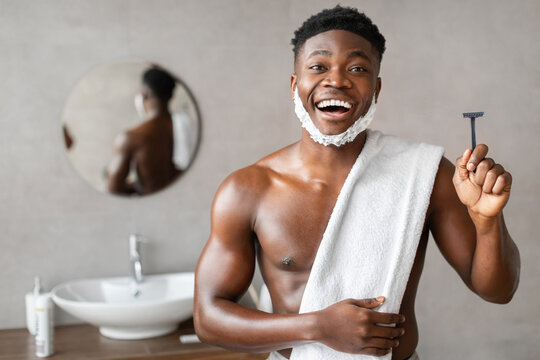 An excited Black man stands in a stylish bathroom, smiling while holding a safety razor. His face is covered in shaving foam, showcasing his grooming routine at home. - Powered by Adobe