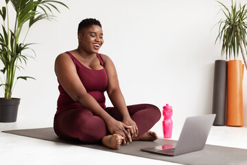 Joyful young black lady in sportswear sits on a fitness mat, smiling while attending an online yoga class. Bright home interior creates a welcoming atmosphere for her workout routine.