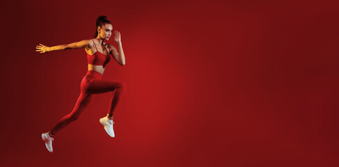 A young woman in athletic wear leaps forward with determination against a solid red backdrop. Her...