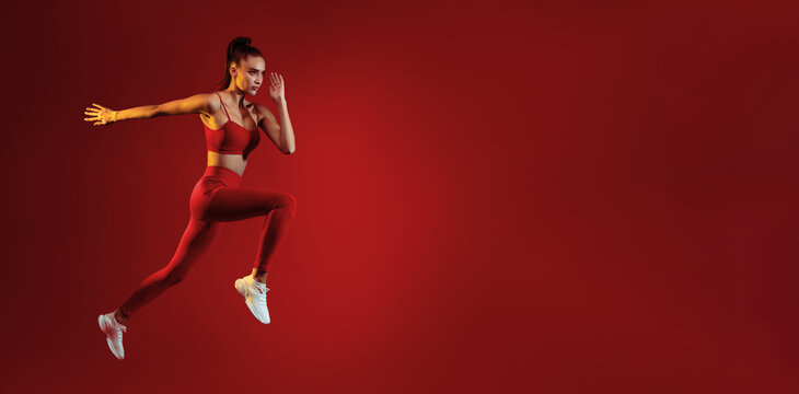A young woman in athletic wear leaps forward with determination against a solid red backdrop. Her focus and movement highlight the energy and strength of her workout. - Powered by Adobe