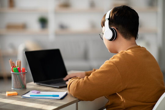 A chubby Chinese boy sits at a desk in his home, wearing a headset and focused on an empty laptop screen. He appears to be engaged in a video conference with a tutor or teacher. - Powered by Adobe