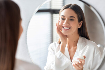 In a bright bathroom, a young woman applies moisturizing face cream while looking at her reflection. She smiles happily, enjoying her beauty routine and self-care moment at home.