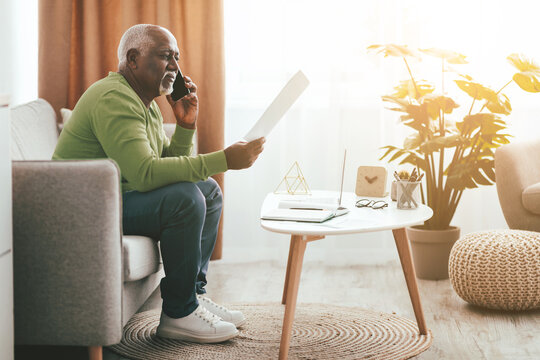 A senior man in a green sweater is seated on a sofa, engaged in a phone conversation. He holds a document and sits beside a coffee table with decorative items in a bright, cozy living room. - Powered by Adobe