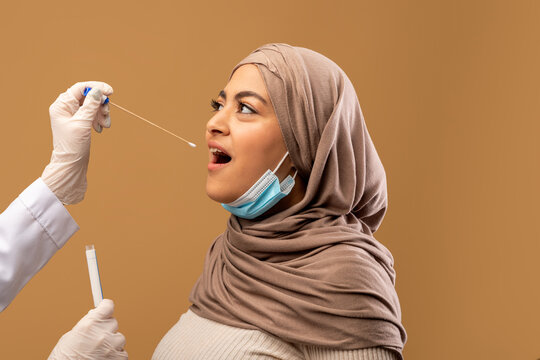 Muslim woman getting PCR oral coronavirus swab test, female doctor in protective medical costume making covid-19 swab test from throat for sick patient, closeup