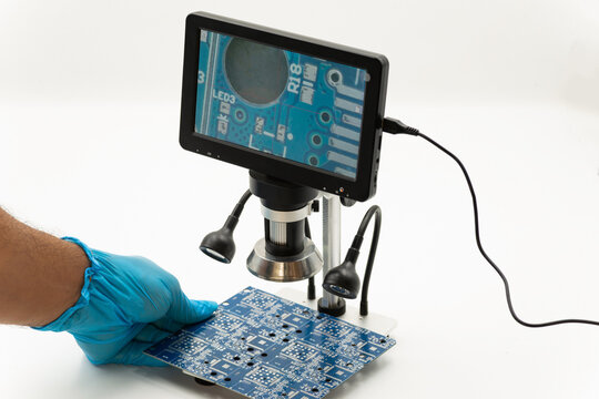 Technician wearing gloves examines a printed circuit board under a digital microscope for quality control, precision inspection, and microelectronic research in a technology laboratory.