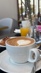 A cozy morning scene featuring a creamy cappuccino with delicate heart-shaped latte art, served alongside pastries and orange juice. The inviting cafe atmosphere promises a perfect start to the day.