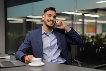 In a modern office, a cheerful arab entrepreneur sits at his worktable, talking on his cell phone. He enjoys a cup of coffee while looking at the space around him, reflecting new business ideas.