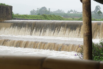 a dam with a fast flowing water, the brown water indicates a strong current due to high rainfall, a sluice gate with a stone and iron structure with a misty mountain background