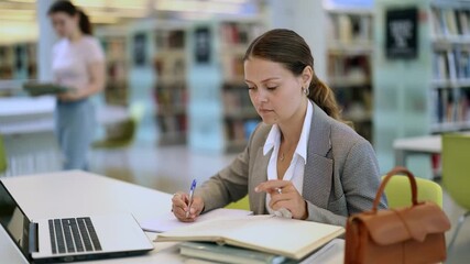 Young female student reading books and working on laptop in library - Powered by Adobe