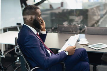 Handsome african american manager having phone conversation with personal assistant, reading papers, checking financial or marketing report, sitting at table at modern office, copy space, side view