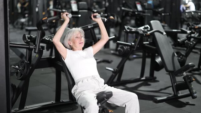 During training session in mini group of athletes, senior woman exercises on shoulder press machine. Client performs exercises, combines dynamic and static loads on body.