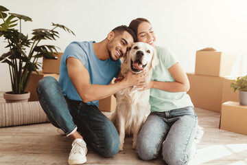 A couple smiles and embraces their golden retriever in a spacious room. Moving boxes and plants are...
