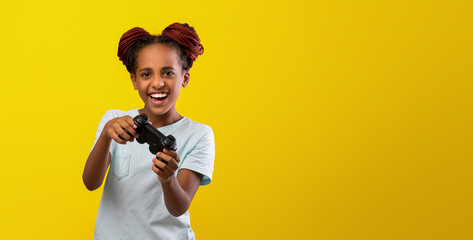 A young girl with braided hair is playing a video game. She is smiling widely, showing her enjoyment. The bright yellow background adds to the cheerful atmosphere of the scene.