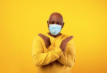 Elderly black man wearing a surgical mask makes a stop gesture with his hands against a bright orange background. He displays a firm stance against the coronavirus and promotes facial protection.