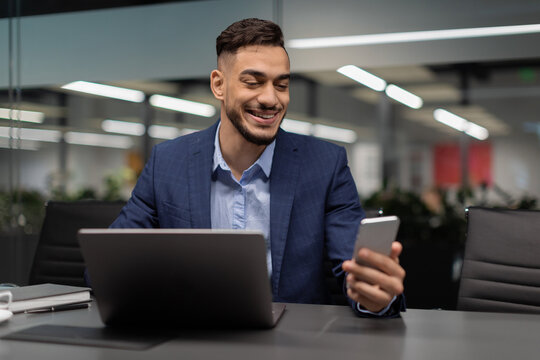 Young Arabic businessman enjoys checking a mobile app while sitting at his workspace. He is focused on his smartphone and laptop in a contemporary office environment.