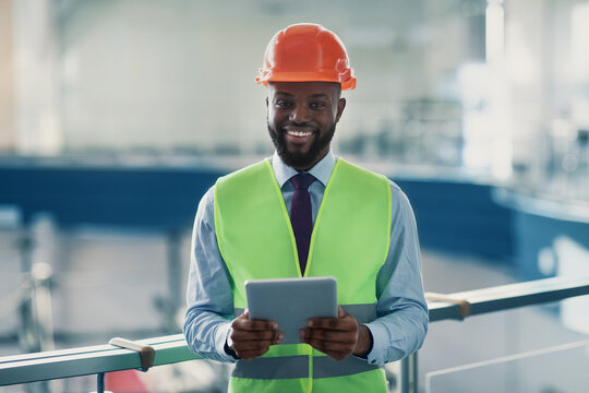 Smiling handsome african american young man in safety vest and helmet civil engineer or architect with digital tablet checking construction site, using modern technologies, copy space