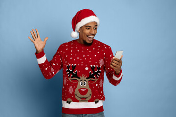 A young man wearing a festive red sweater with a reindeer design and a Santa hat joyfully waves while checking his phone. The background is a soft blue.