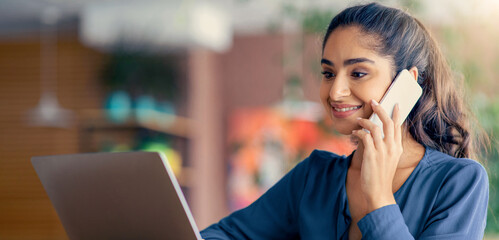 A young woman is smiling as she talks on the phone while using her laptop in a modern office. Colorful decor in the background adds to the vibrant atmosphere.