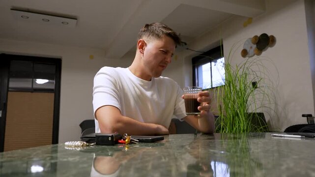A man reacts with a disgusted facial expression while smelling and tasting burnt coffee in a modern kitchen, showing displeasure, surprise, and a strong negative reaction