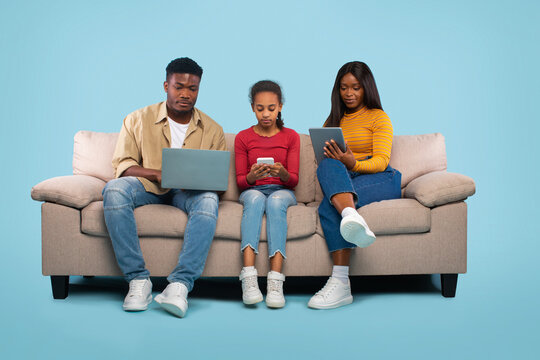 A family is sitting on a light-colored couch engaged with their electronic devices. The father uses a laptop, the mother browses on a tablet, and their daughter plays with a smartphone.