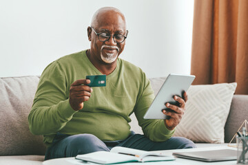 An elderly man sits comfortably on a sofa, smiling as he uses a tablet and holds a credit card in his hand. Natural light fills the room, creating a warm atmosphere.