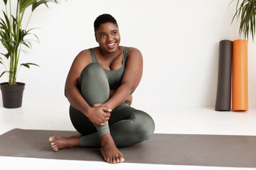 A young, attractive African American lady sits on a yoga mat with a smile. She is wearing tight sportswear and engages with the space. The setting is light and encouraging, perfect for yoga practice.