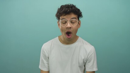 Man yawns showing mouth and glasses in studio setting on teal background under subtle soft light; fatigue.