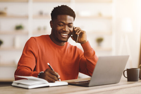 A man in a red sweater smiles on a phone call while taking notes in a notebook. His laptop sits open on a table, creating a productive home office setting filled with natural light. - Powered by Adobe