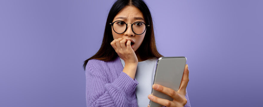 A young woman with glasses looks shocked as she checks her phone. She is biting her nails and wearing a purple sweater. The background is a soft purple, emphasizing her expression of surprise.