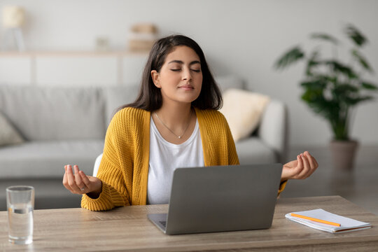A calm arab woman with closed eyes meditates in front of her laptop at a desk, promoting workplace stress management. She embodies peace while freelancing in a cozy home office.