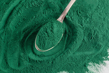 A metal spoon with natural organic green algae powder rests on the scattered spirulina powder. top...