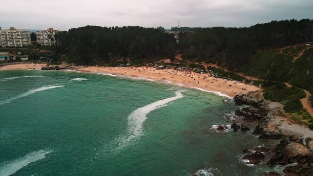 Backward drone aerial of Canelo Beach, Algarrobo, Chile, turquoise Pacific waters with tourists enjoying summer vacation on sandy shore