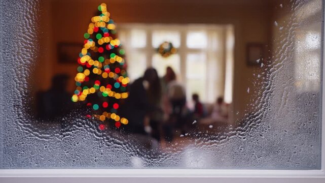 Frost covered window pane from inside a cozy room with warm blurry shapes of a family and christmas tree, concept of warm family contrast to cold winter
