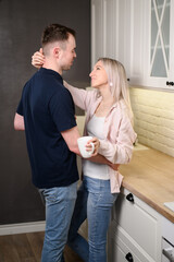 Affectionate partners communicate in cozy atmosphere of kitchen, standing next to each other, hugging, gazing lovingly into each other's eyes, holding mug. Hugs, romance, happy married, caring, trust.