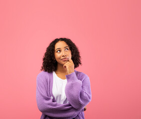 A thoughtful African American woman touches her chin as she considers an offer. She appears uncertain while gazing into empty space, surrounded by a soft pink background.