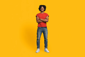 A smiling young man with curly hair is standing with arms crossed. He wears a red shirt and jeans,...